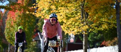 Two riders on a country road pass by rows of colorful trees and a white picket fence.